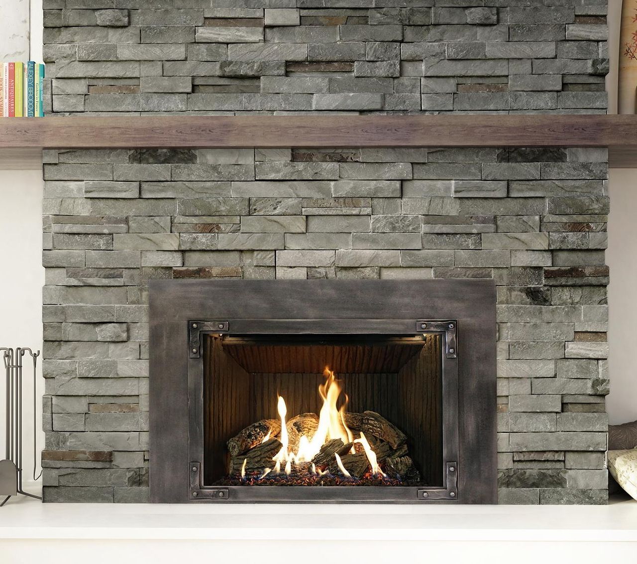 A stone fireplace with a lit fire inside. A dark metal frame surrounds the firebox, and a wooden shelf holds books above.