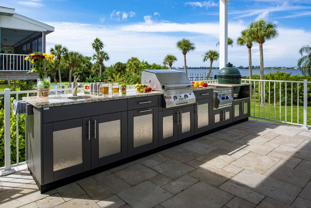 Outdoor grilling kitchen on a sunny patio with a gray cabinet, a grill, a Big Green Egg, and a sink. Palm trees and water in the background.