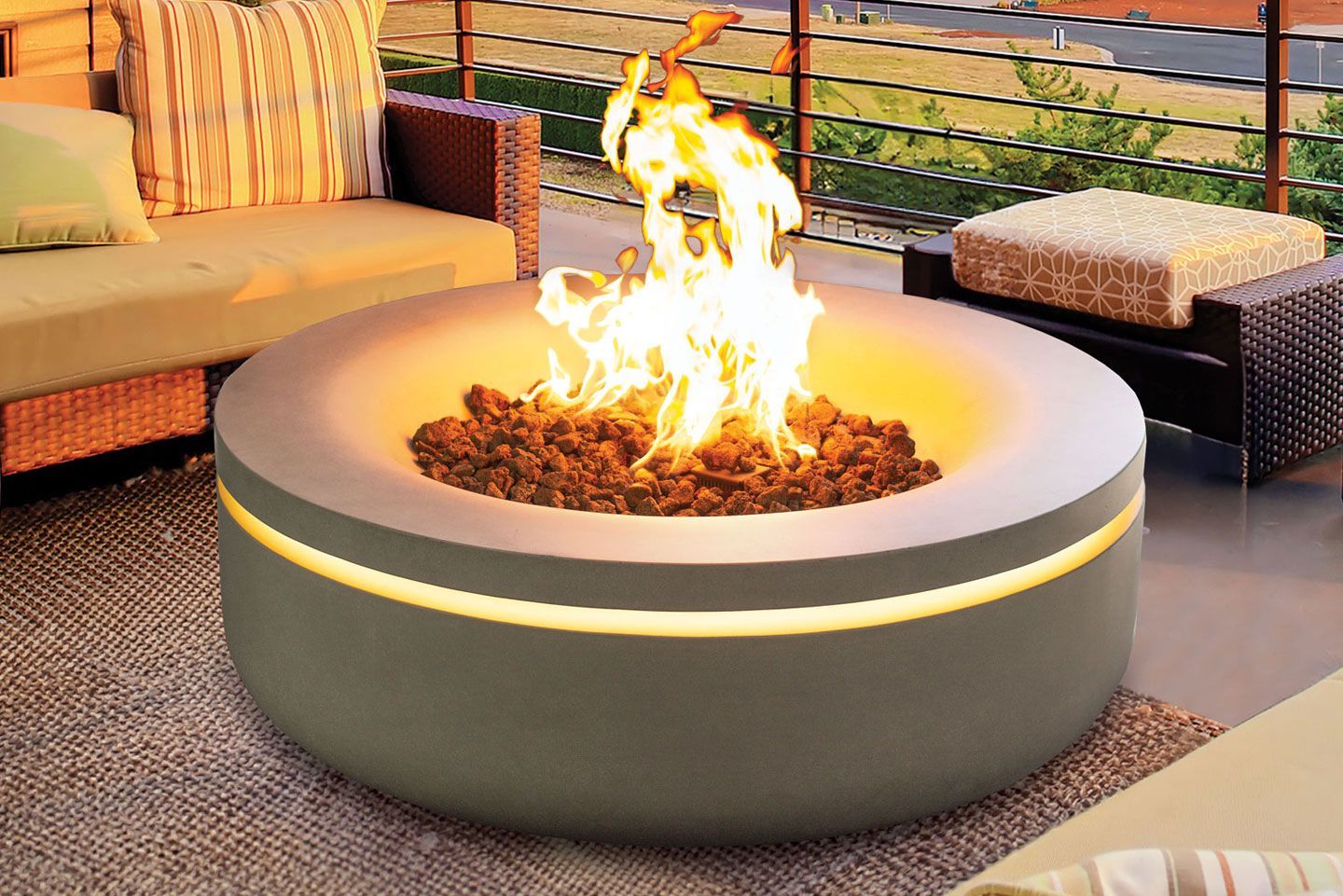 Outdoor seating arrangement with a fire pit table and wicker furniture. Gray cushions, flames, and a wooden deck against a white picket fence and trees.
