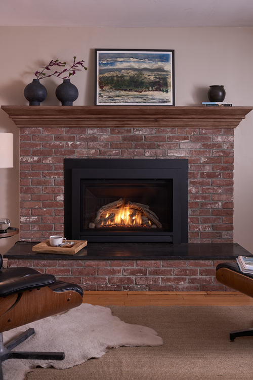 Brick fireplace with a black firebox, topped with a wooden mantel displaying art and vases. A burning fire glows inside the box.