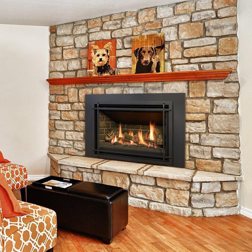 Corner fireplace with stone surround, wooden mantel, and two dog portraits. Two patterned armchairs and a black ottoman are nearby.
