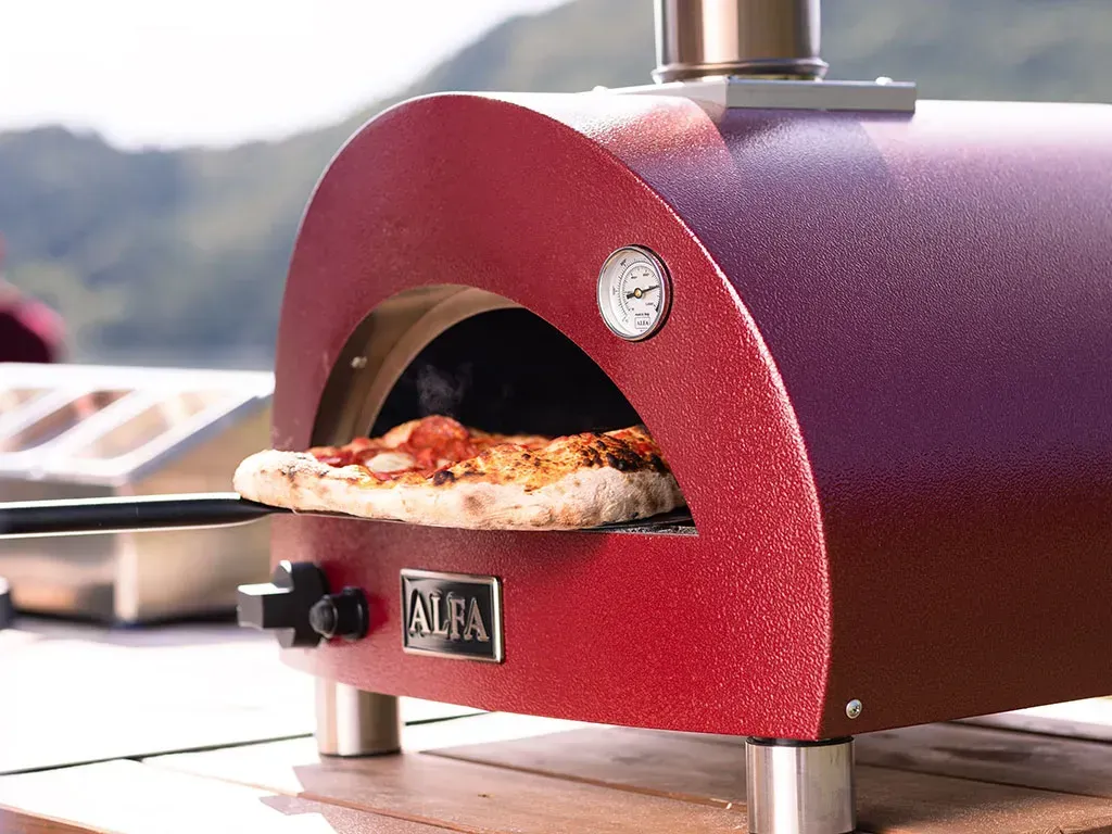 A red outdoor pizza oven baking a pizza. The oven is on a wooden surface, with a stainless steel chimney and legs.
