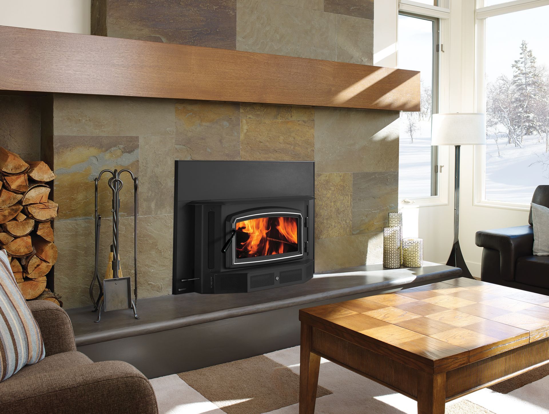 Fireplace with a burning fire, brown stone surround, and wood mantle. Fireplace tools are arranged on the hearth, and a stack of firewood is next to a brown armchair.