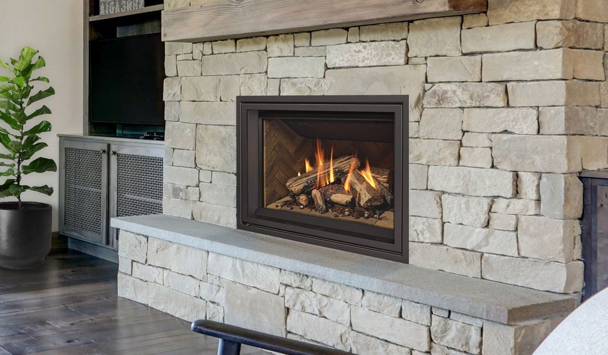 Gas fireplace with burning flames and wood logs set in a stone wall, with a wooden mantel and a living room setting.