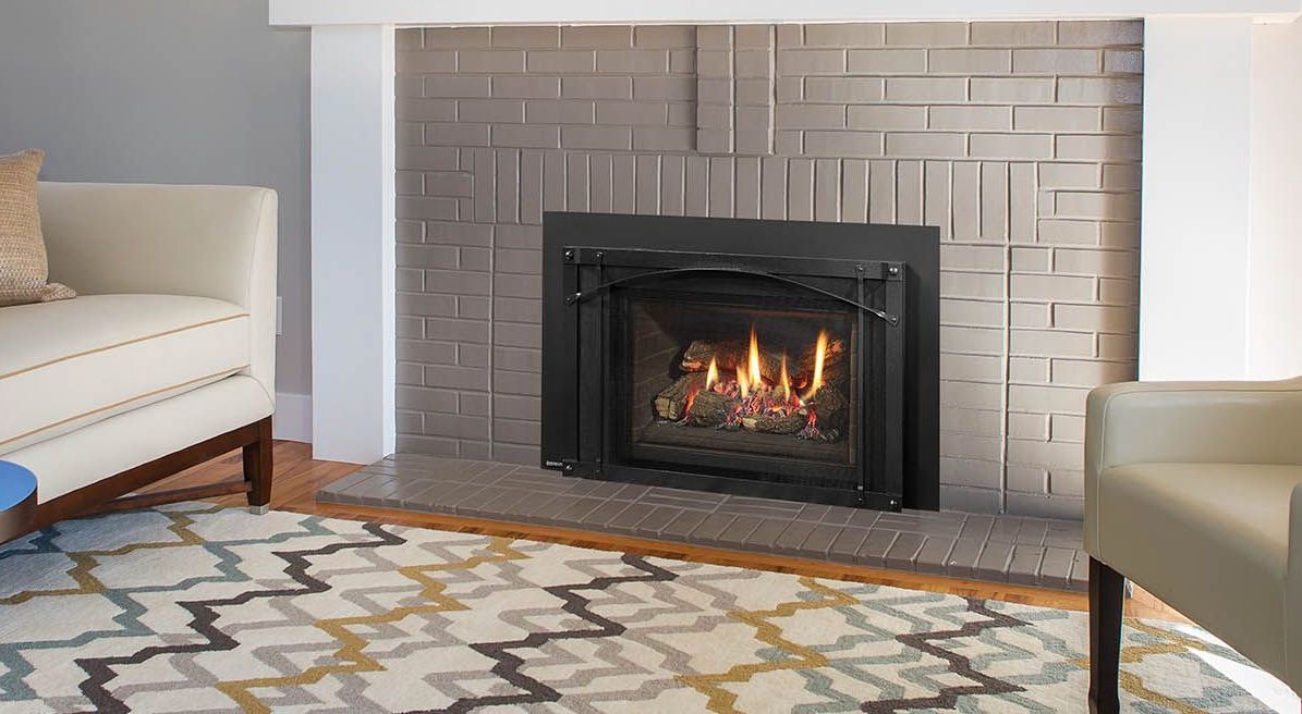 A living room with a fireplace, lit with a flame. The brick surround is gray, and the room has a patterned rug and cream-colored furniture.