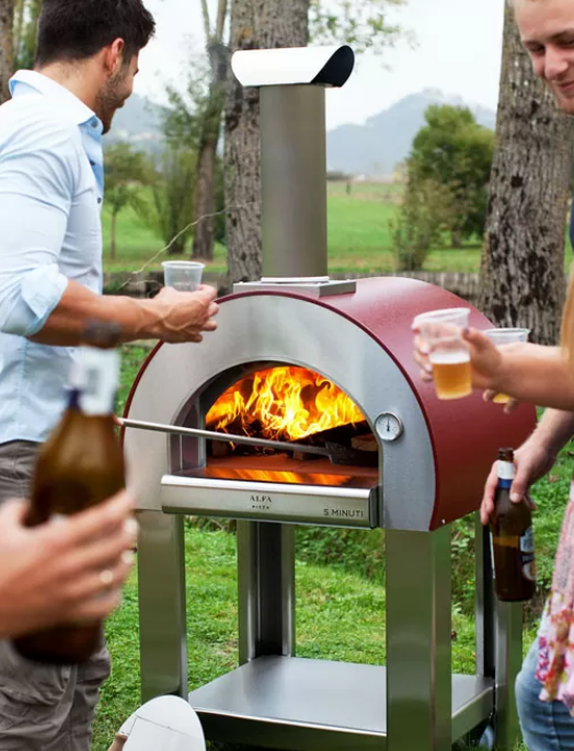 People around a red outdoor pizza oven with flames, holding drinks in a park setting.