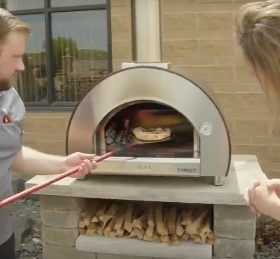 A man uses a long tool to maneuver a pizza inside a stainless steel outdoor pizza oven, while a woman watches. The oven sits atop a brick structure.