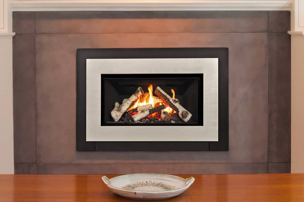 A fireplace with burning logs inside a black and silver frame, set in a brown wall. A decorative bowl sits on a wooden table below.