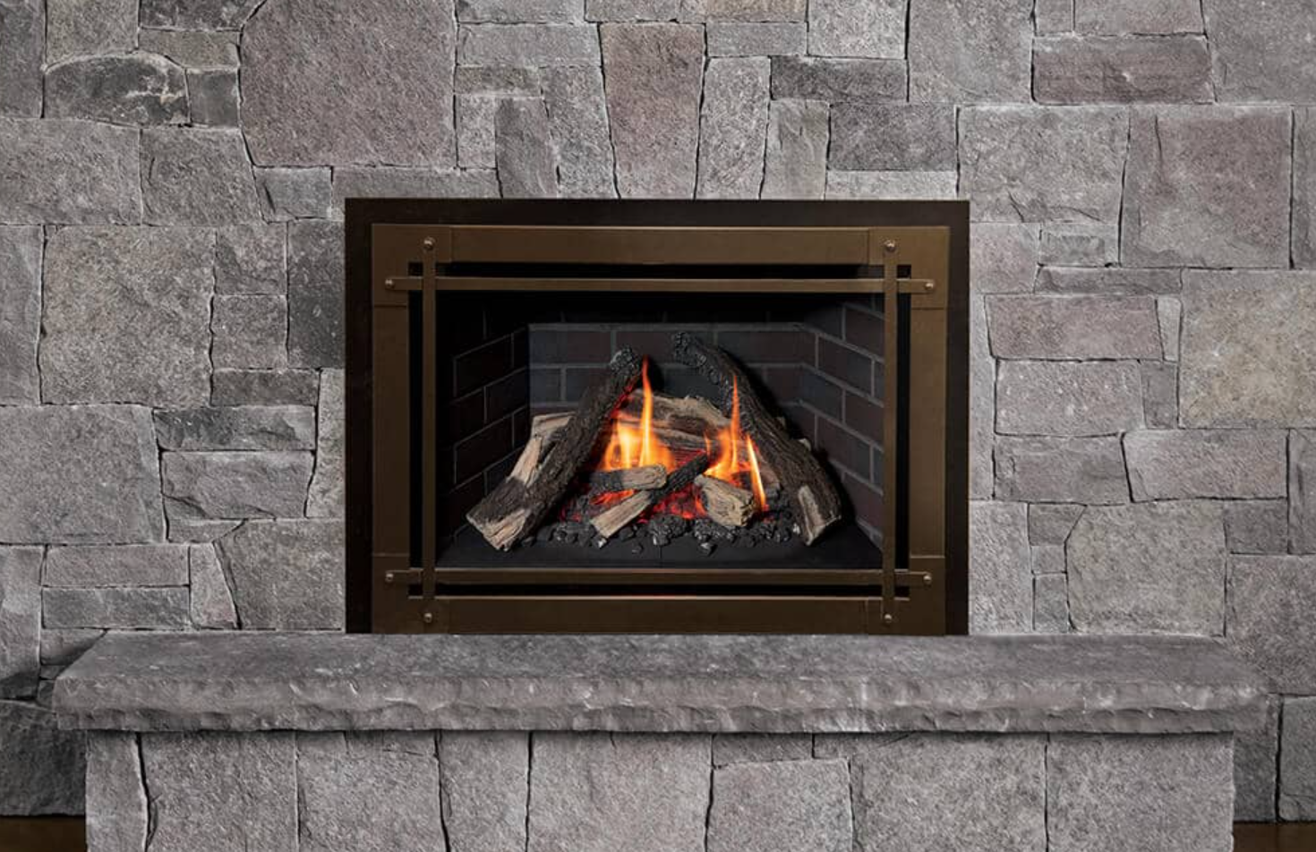 Fireplace with burning logs and a bronze-colored metal frame set against a gray stone wall and hearth.