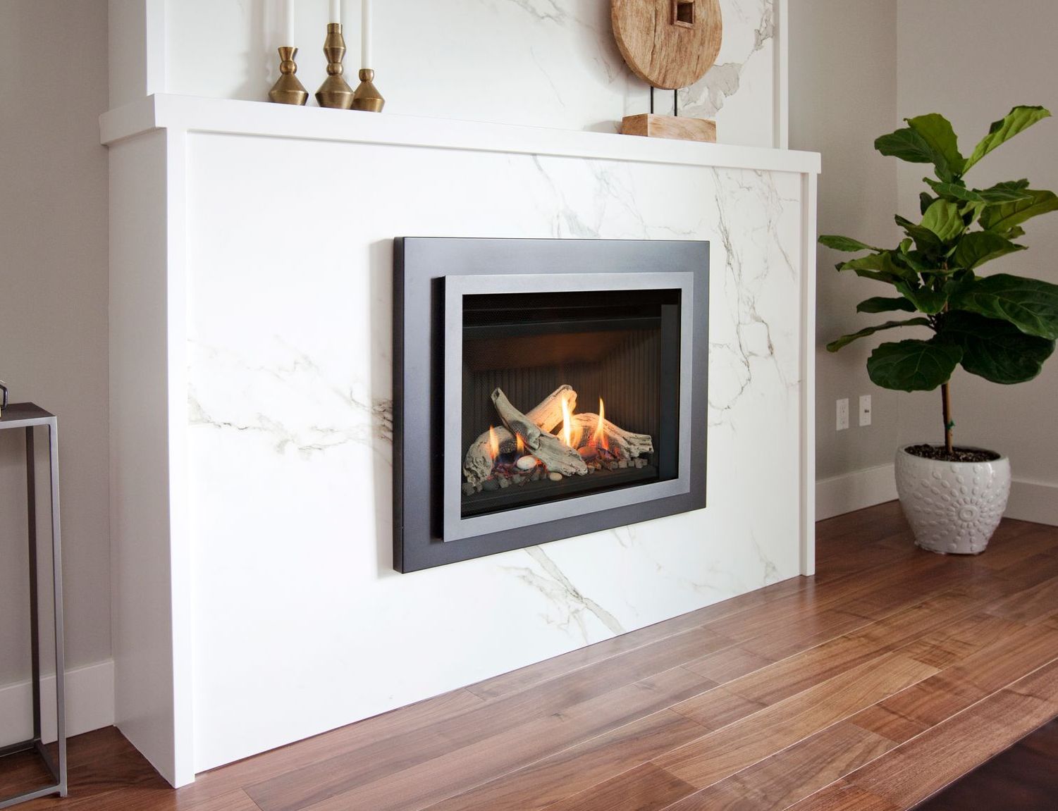 Fireplace built into a white marble wall, with burning flames inside. A potted plant is to the right.