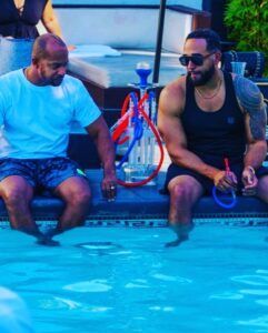 Two men are sitting on the edge of a swimming pool with hookahs.