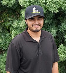 A man wearing a black hat and a black polo shirt is standing in front of a bush.