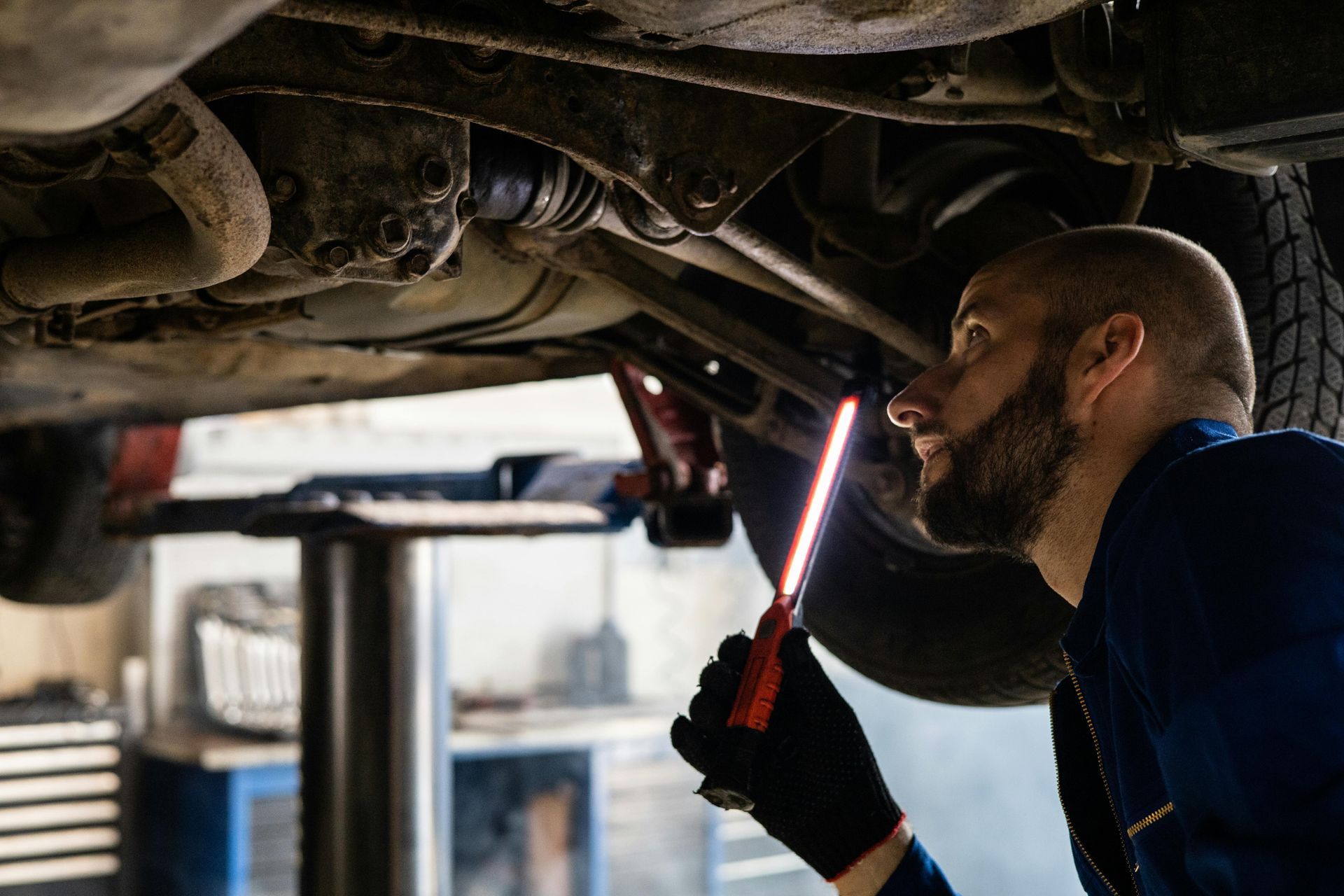 A man is working under a car in a garage with a flashlight.