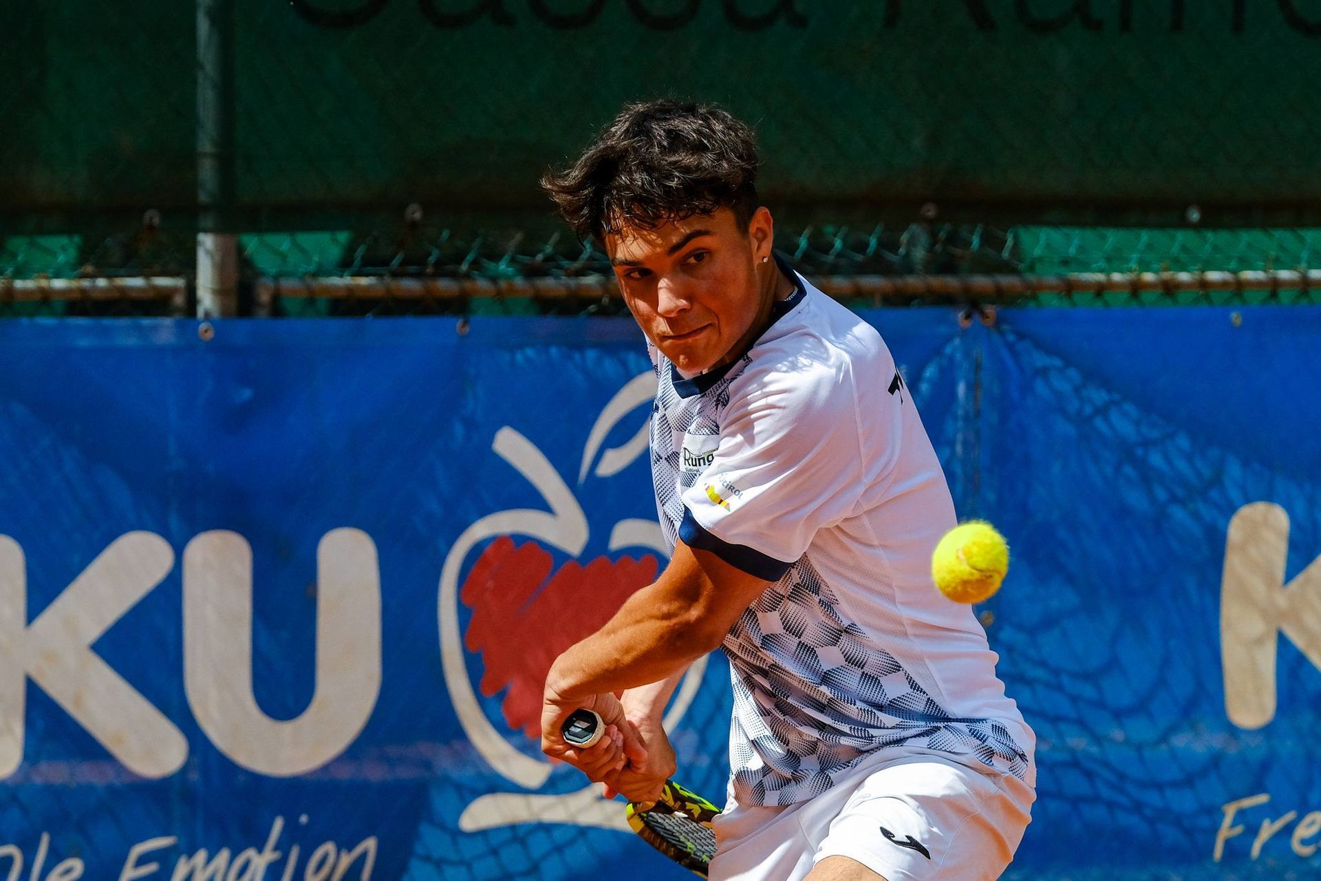 A man is playing tennis in front of a banner that says ku