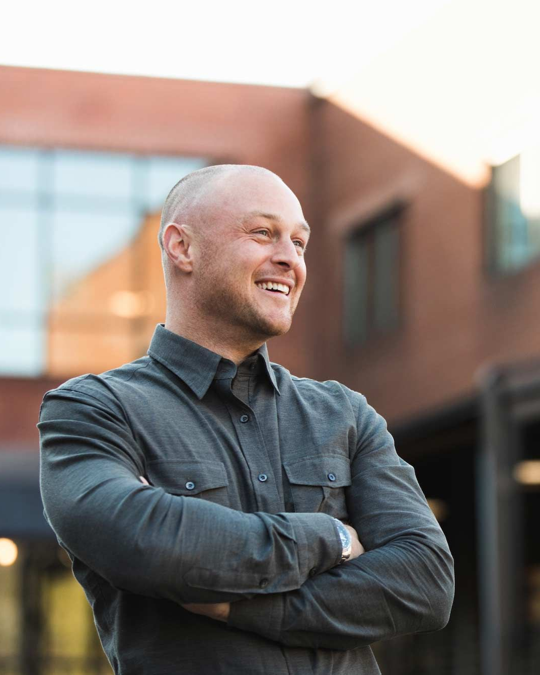 Man with arms crossed smiling, wearing a dark button-down shirt outdoors in front of a brick building.