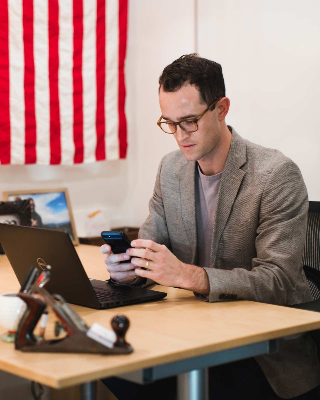 Man in glasses looks at phone, sitting at desk with laptop, American flag in background.