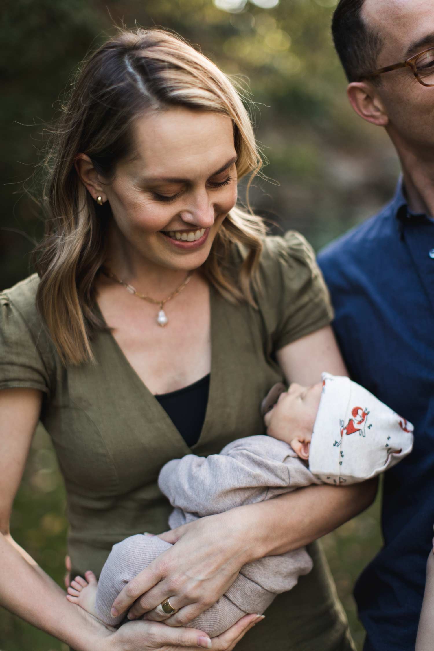 Woman in olive green shirt smiles, holding sleeping baby in arms. Man partly visible. Outdoors.