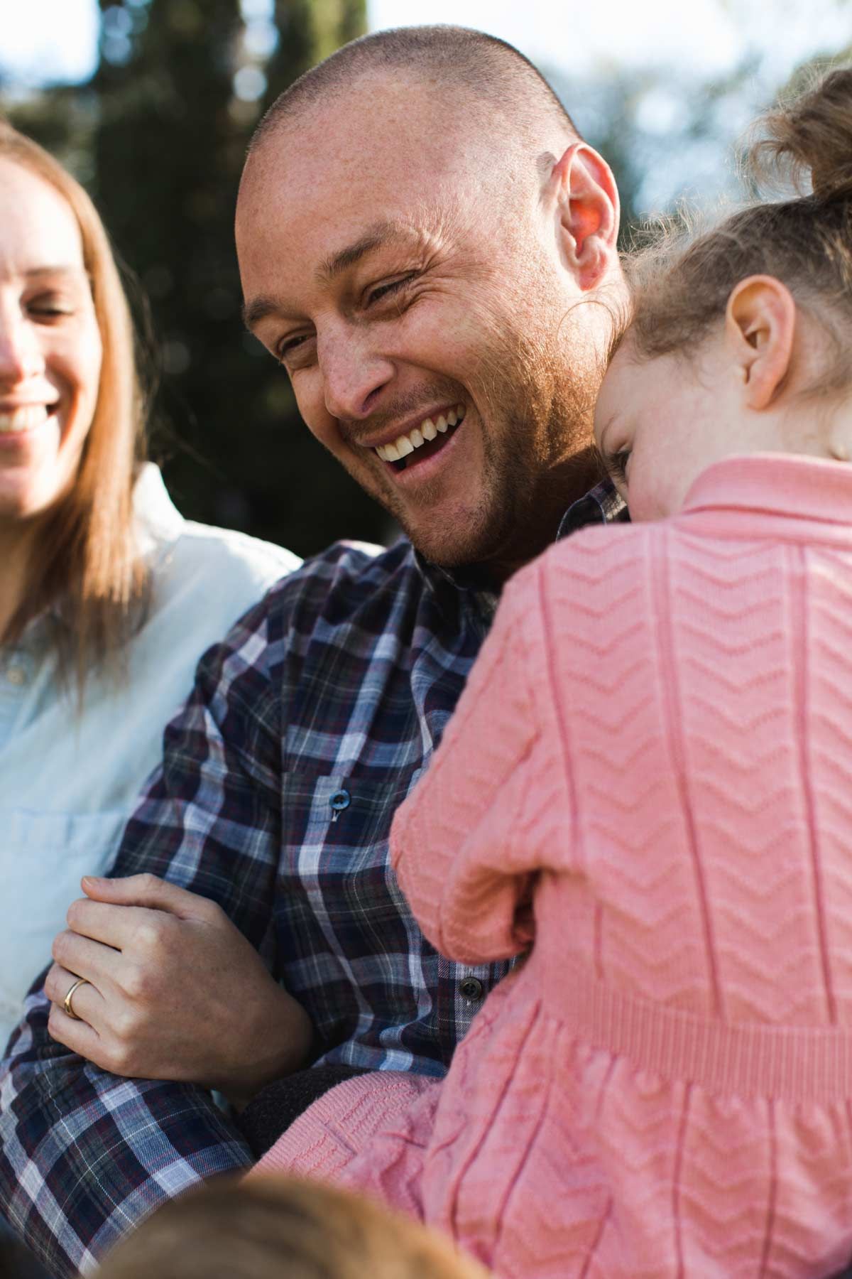 Man laughing, holding a child in pink sweater. Woman in light blue shirt smiles nearby. Outdoors.