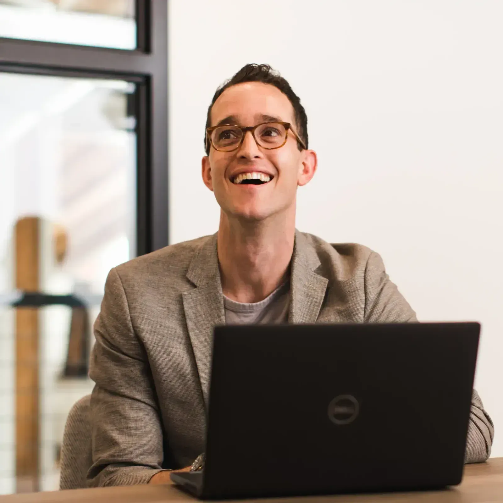 Man in glasses smiles, looking up while sitting at a table with laptop. Office setting.