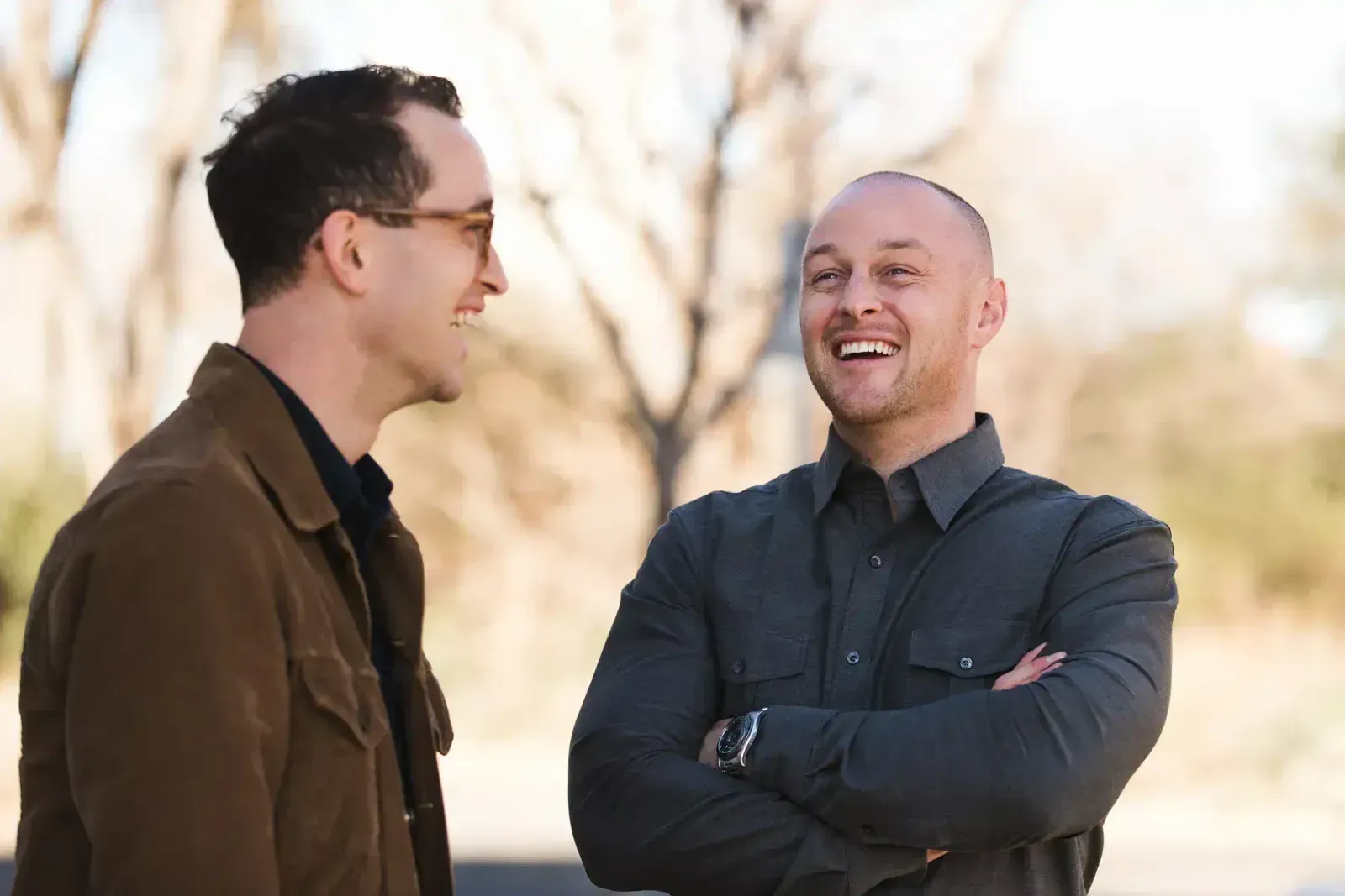 Two men laughing outdoors, one with sunglasses and a brown jacket, the other with arms crossed, wearing a gray shirt.