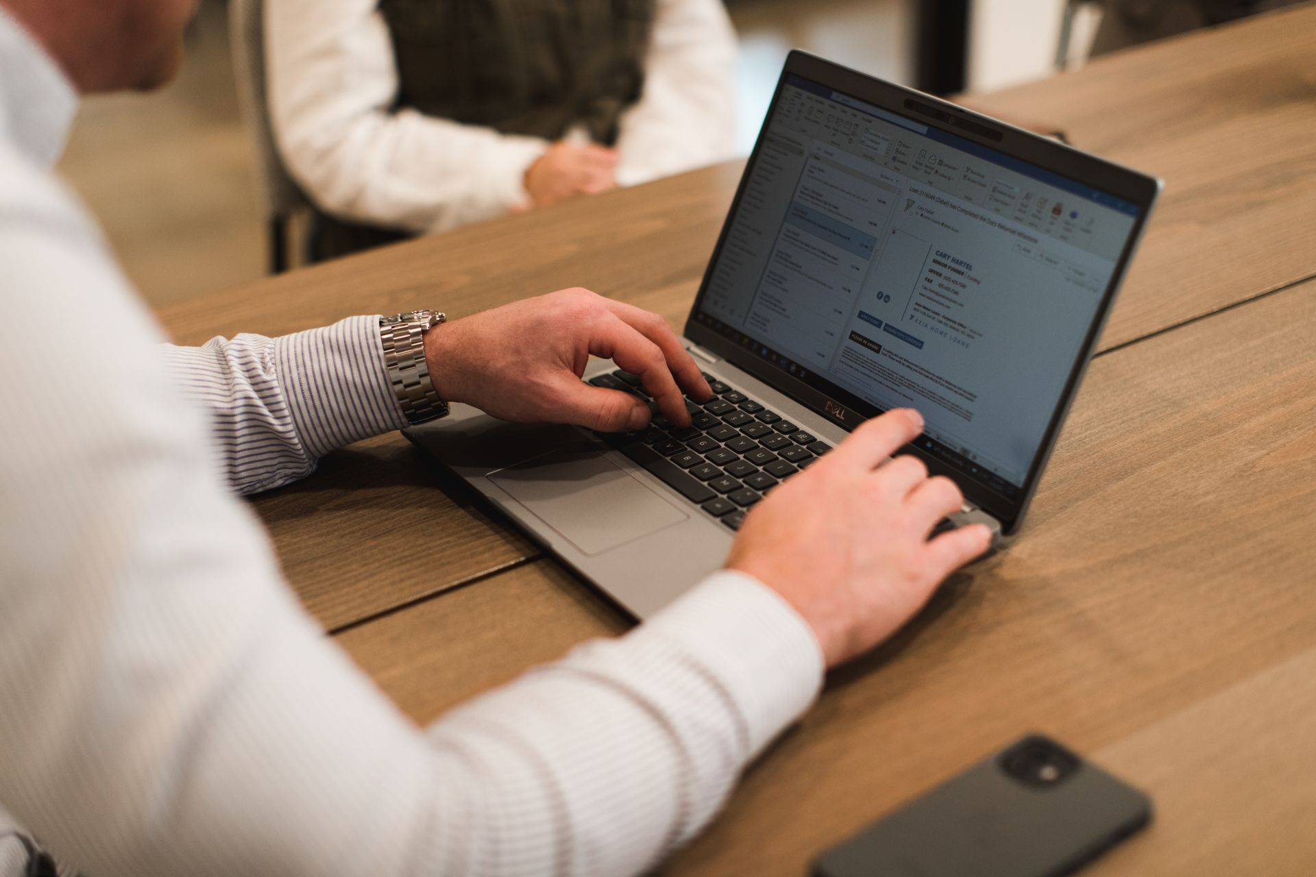 Person typing on laptop at wooden table, with another person out of focus in background, and phone next to laptop.