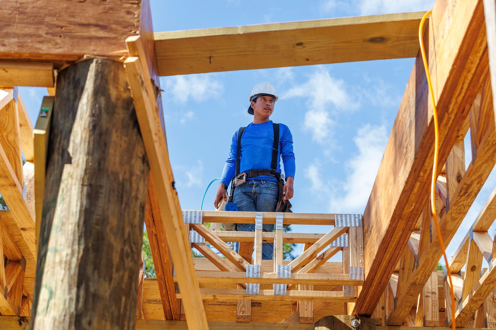 A male construction worker stands on top of the wooden rooftop frame of a house.