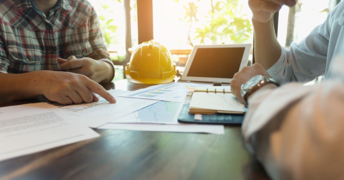 Two people review documents spread out on a table. A yellow hard hat, a tablet, and a notebook are also on the table.