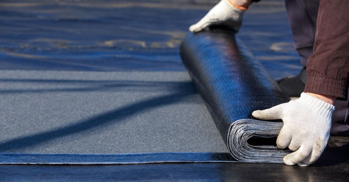 Close-up of a worker's white gloves holding a partially unrolled sheet of black roofing material on a flat roof surface.