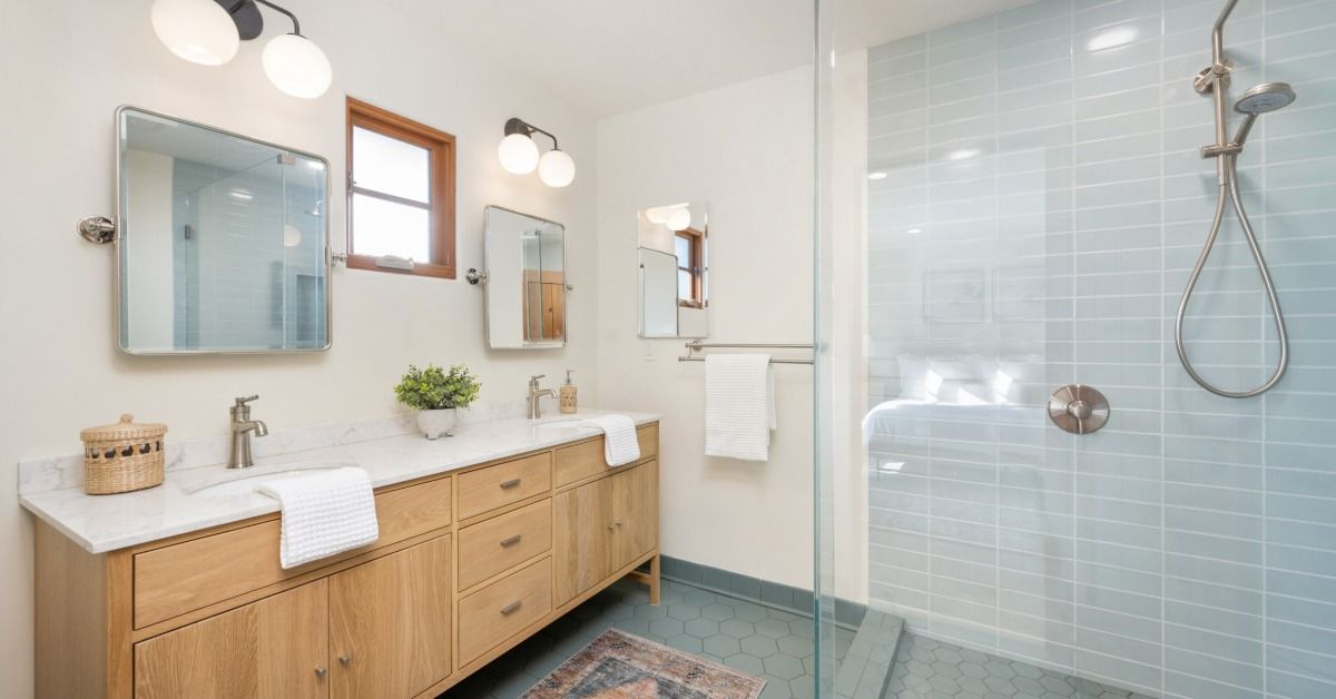 A double-sink wooden vanity is across from a glass-enclosed shower with light blue tiles and a handheld showerhead.