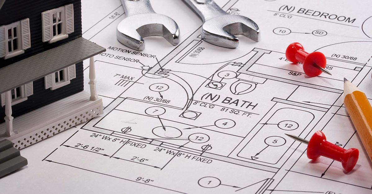 A miniature house model, wrenches, a pencil, and red push pins sit atop a 2D home floor plan of a be