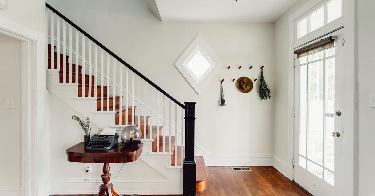 In a bright white entryway, a wooden staircase descends from the left. On the right is a glass-paneled door.
