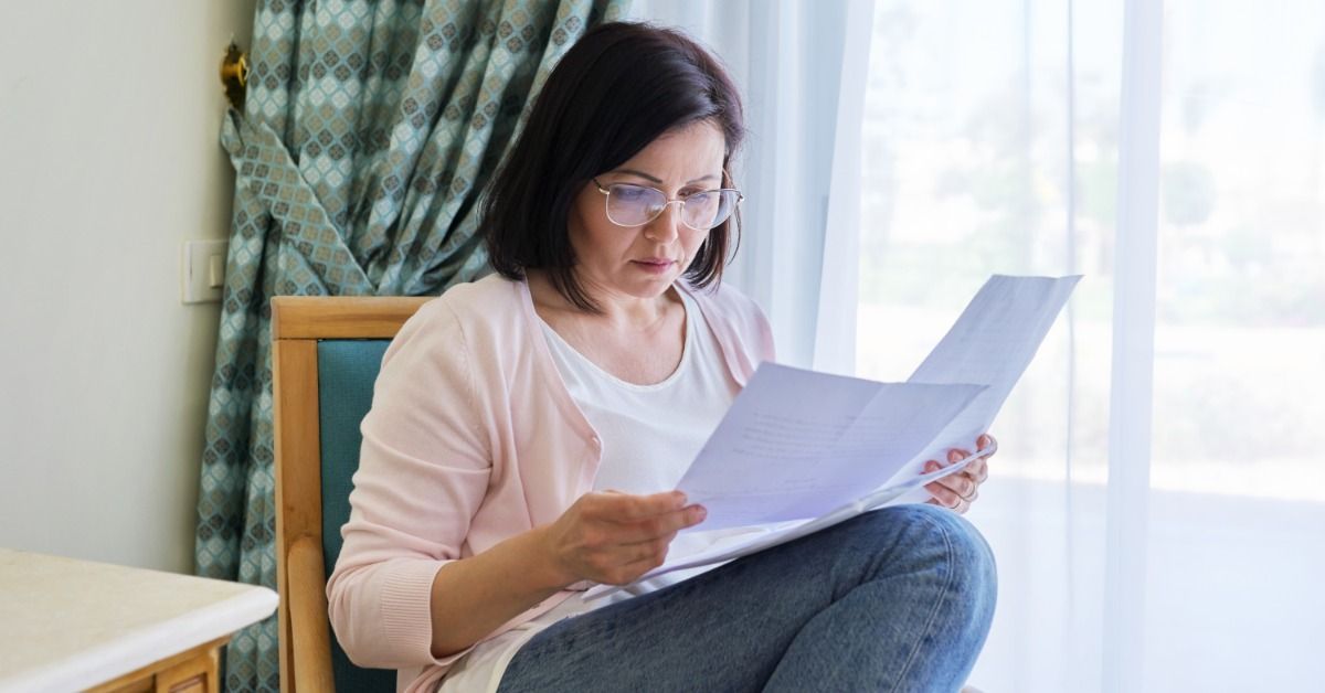 A woman wearing glasses sits in a chair by a large, curtained window. She holds and reads several sheets of paper.