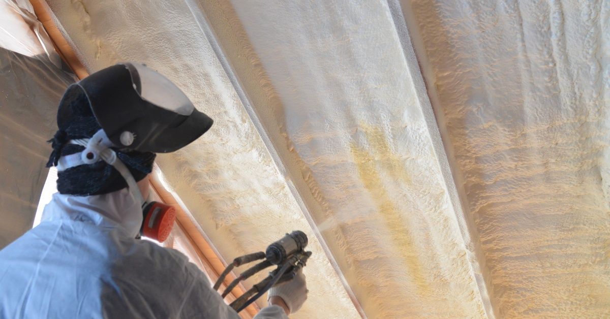 A worker wearing protective gear sprays white foam insulation between ceiling beams using a handheld hose.