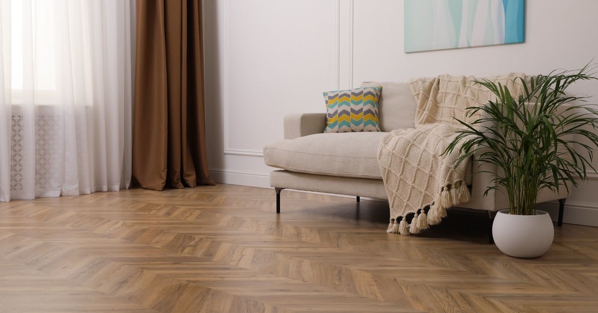 A living room with a light-colored sofa and a potted plant. The floor features a herringbone-pattern
