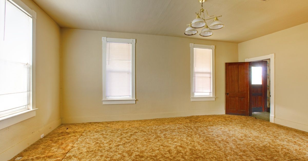 An empty room with beige walls and multiple windows. The old yellow-and-brown carpet has ripples in different places.