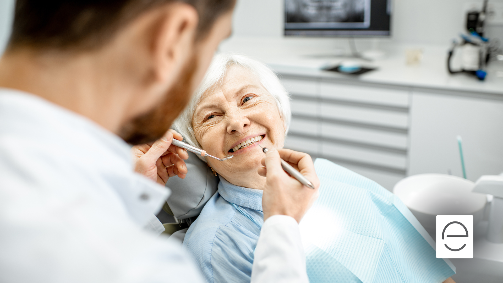 Dentist examining an elderly woman's teeth in a dental clinic. She smiles while he holds instruments.