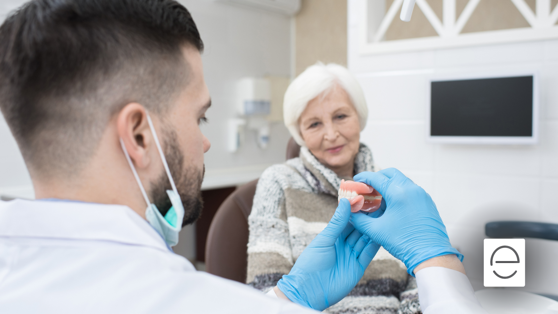 A dentist is holding a denture in front of an elderly woman.