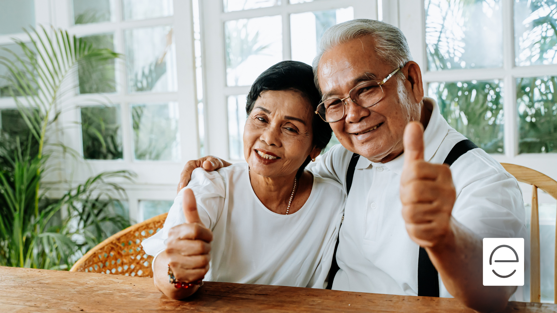 An elderly couple is sitting at a table giving a thumbs up.