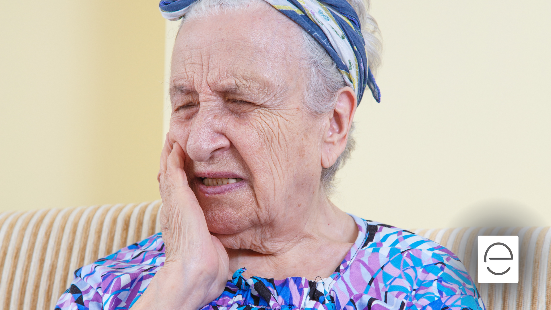 An elderly woman is sitting on a couch with a toothache.