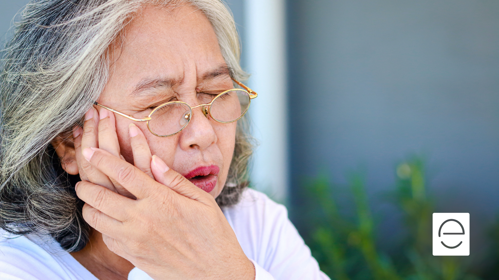 An elderly woman with glasses is holding her mouth in pain.