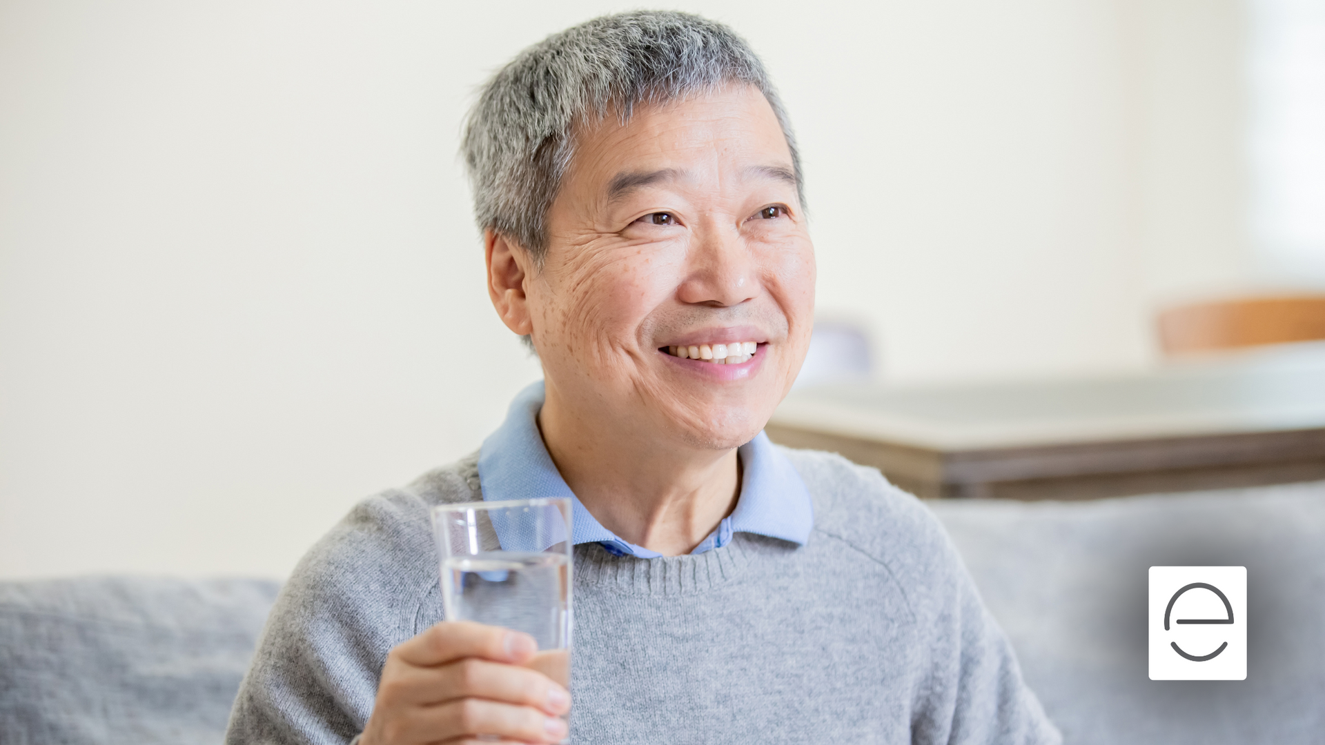 An elderly man is drinking a glass of water while sitting on a couch.