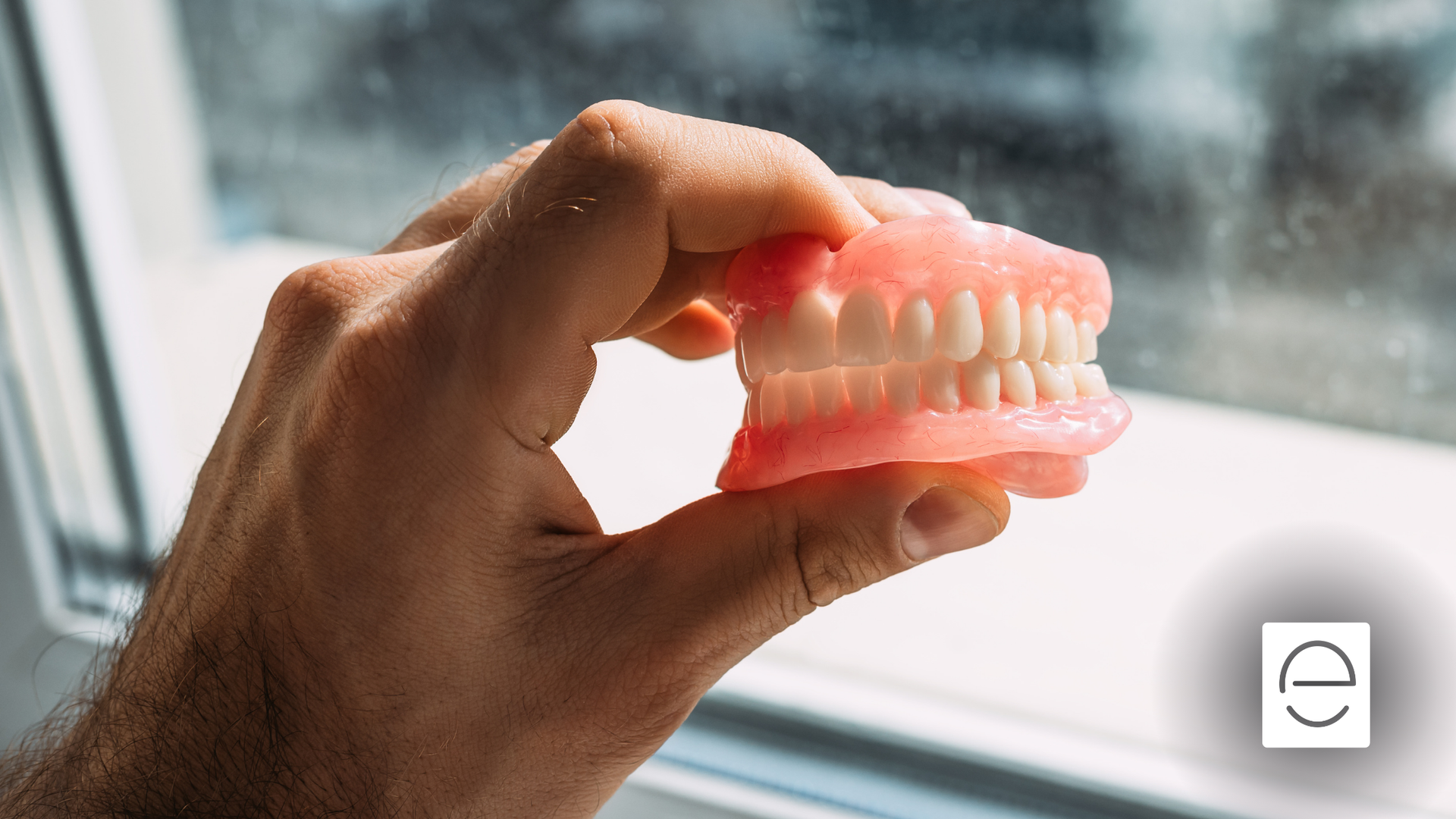 A person is holding a denture in their hand in front of a window.