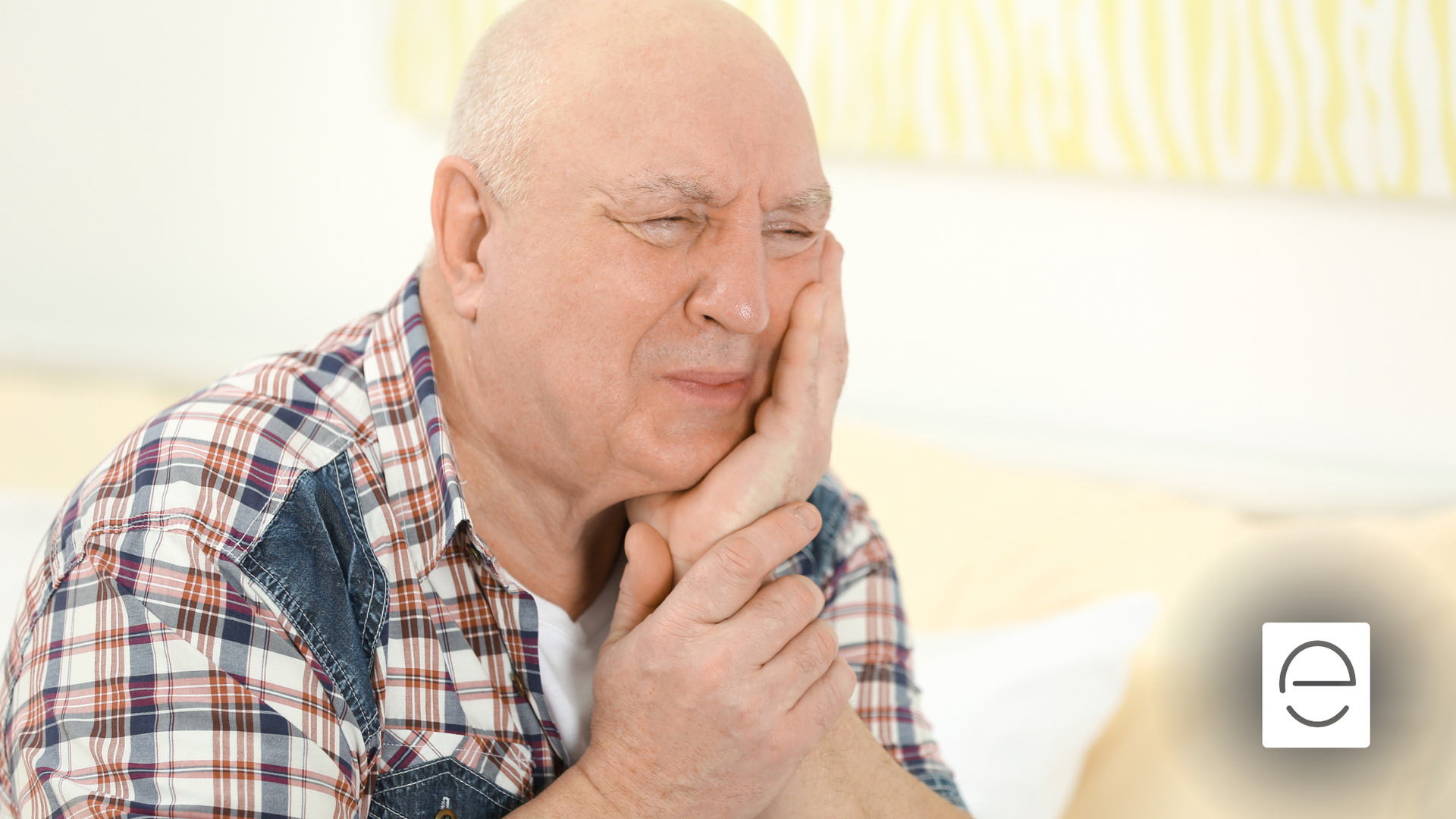 An elderly man is holding his face in pain while sitting on a couch.