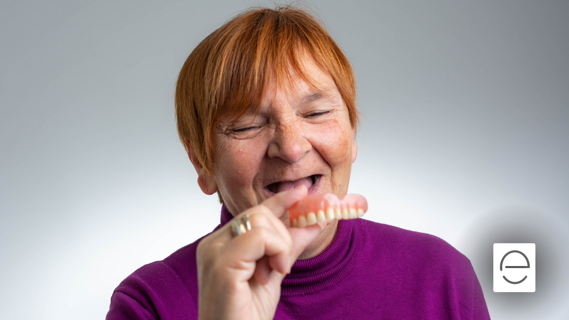 An elderly woman is holding a denture in her mouth.