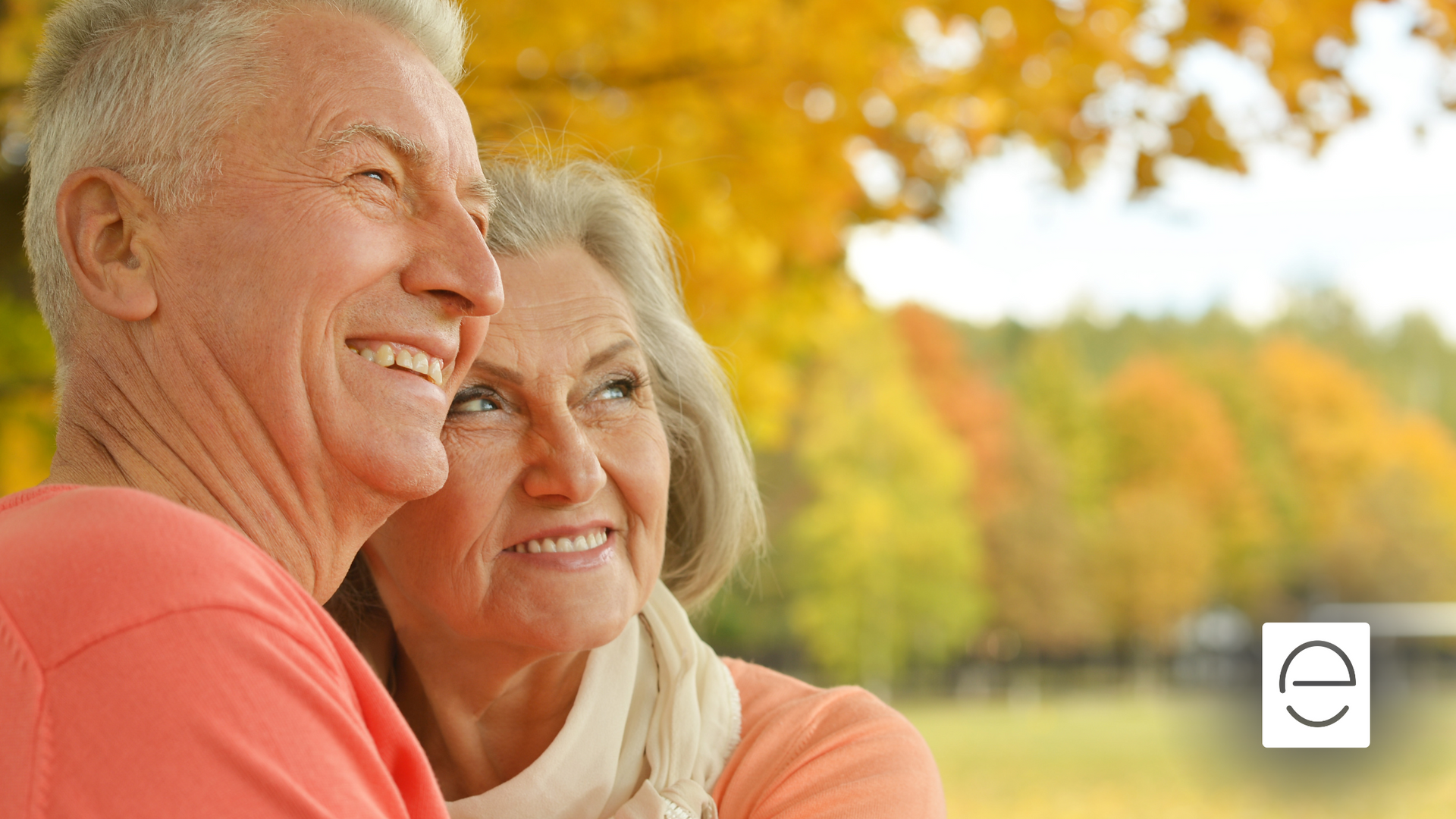 An elderly couple is standing next to each other in a park and smiling.