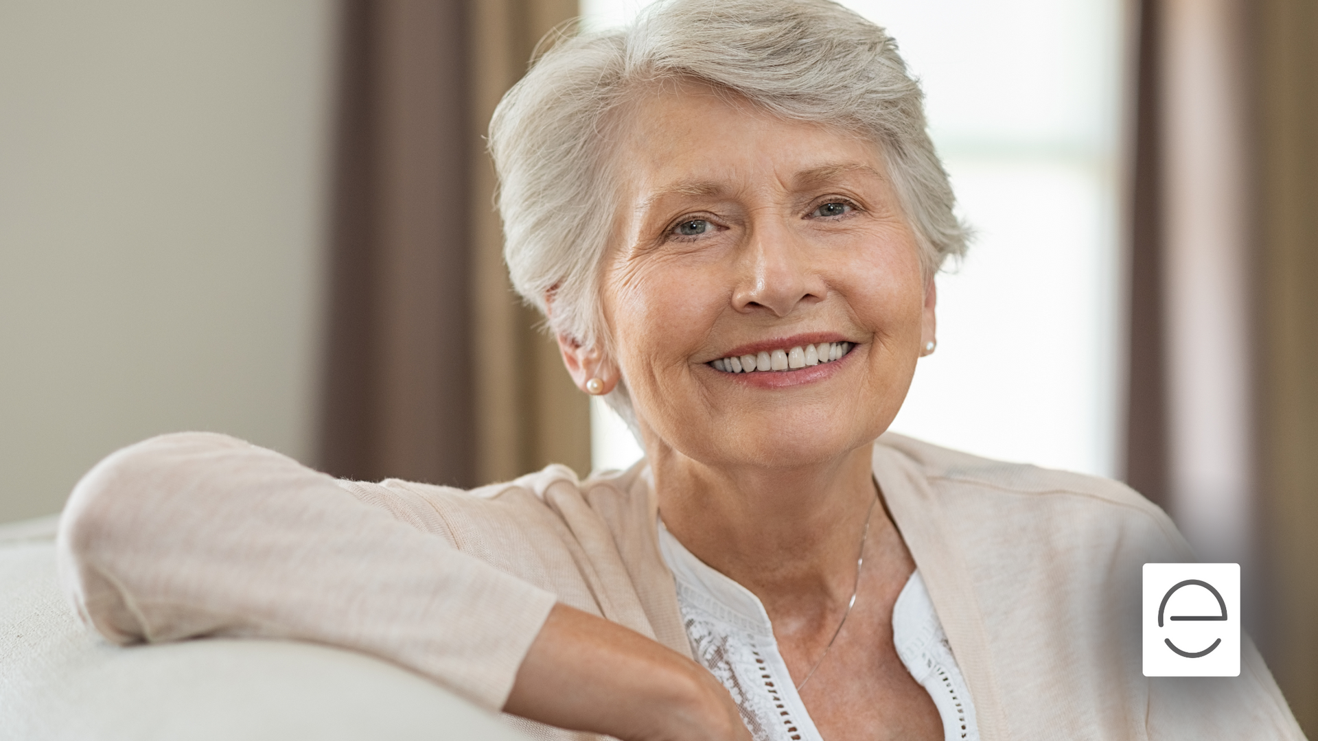 An elderly woman is smiling while sitting on a couch.