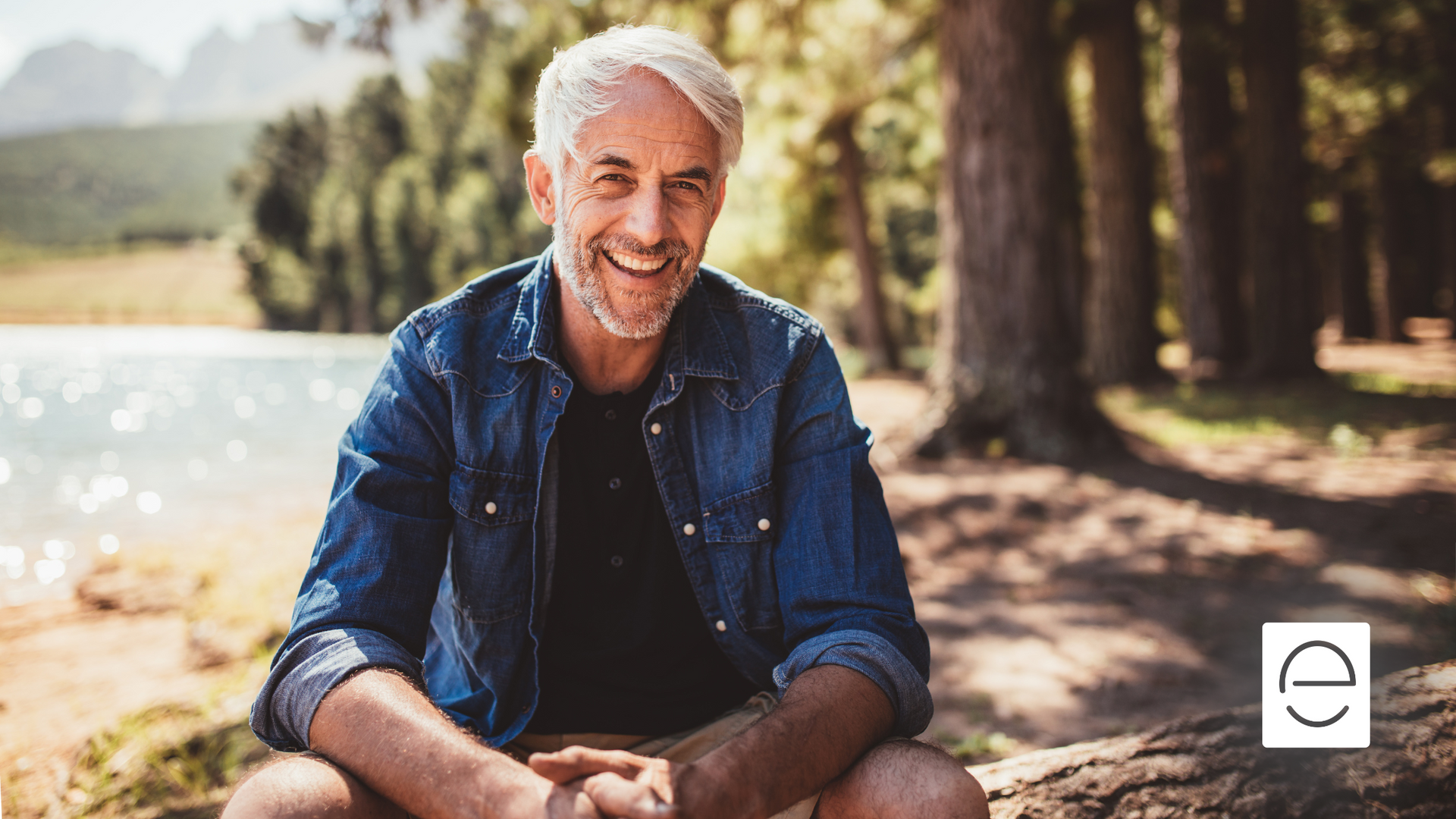 A man is sitting on a rock near a lake and smiling.