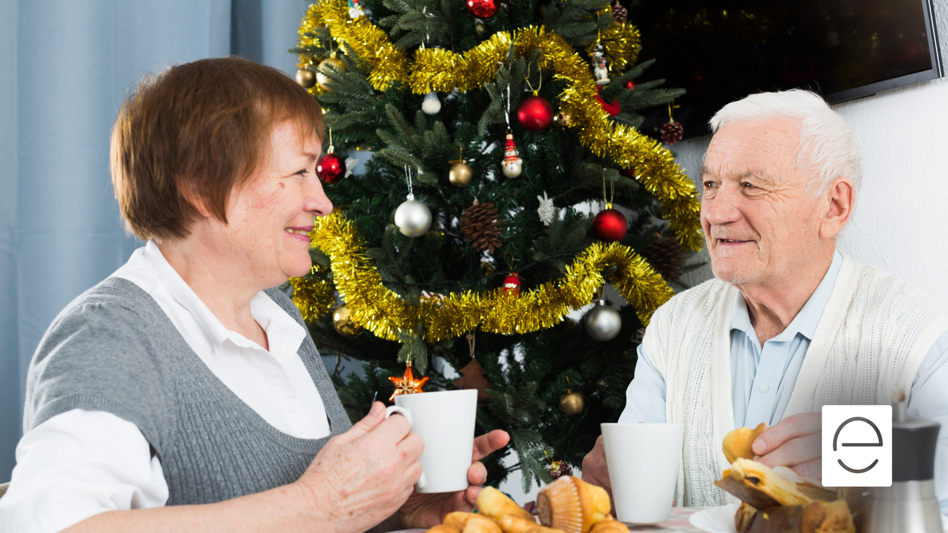 An elderly couple is sitting at a table in front of a christmas tree.