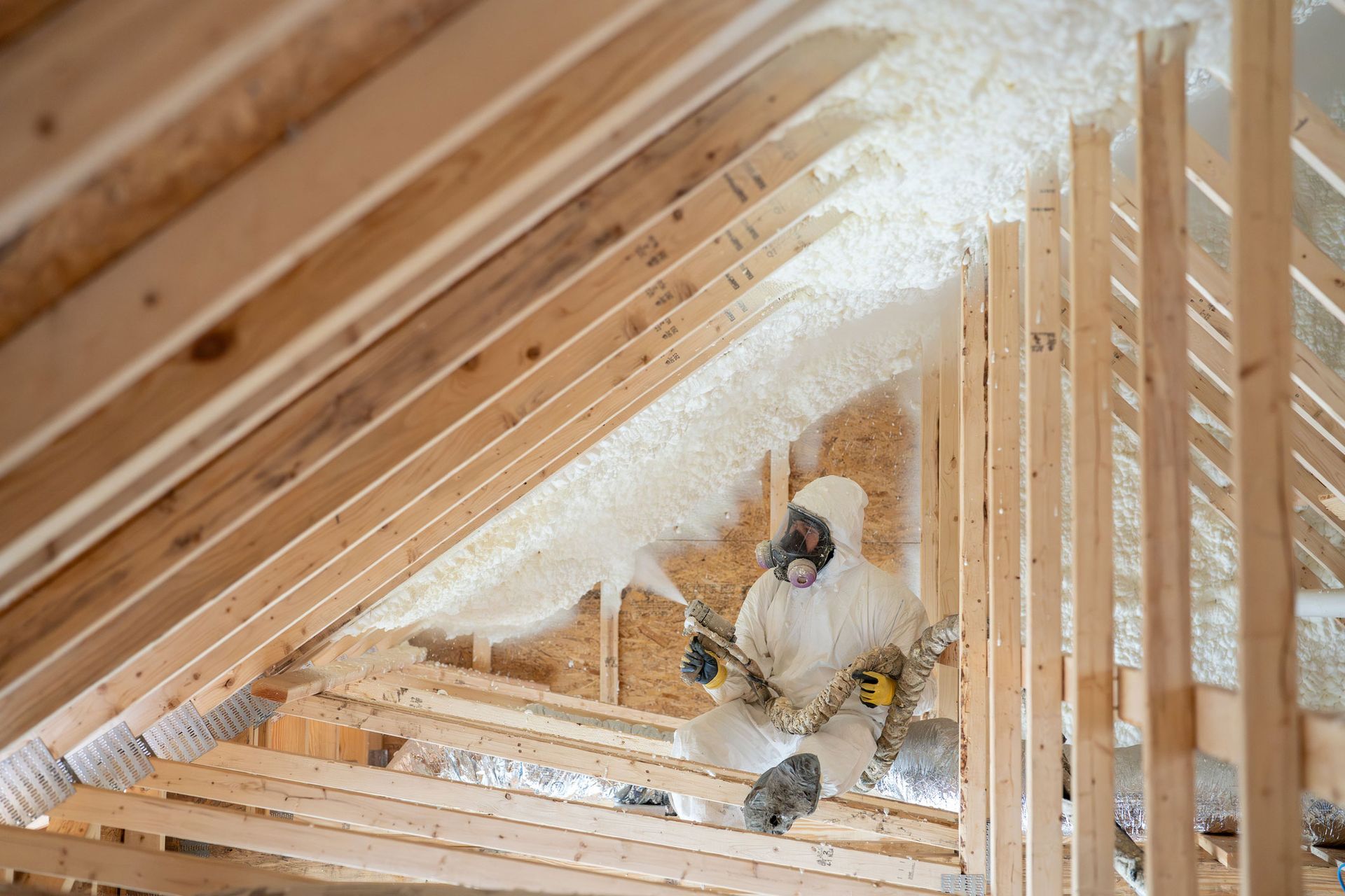 Worker in protective gear spraying foam insulation in an attic.