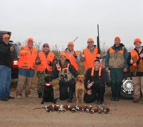 Group of hunters in orange vests with dogs and harvested pheasants, posing outdoors.